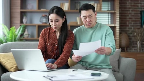 Frustriertes schockiertes asiatisches Ehepaar schaut sich große Stromrechnungen mit einem Laptop an, der zu Hause sitzt.