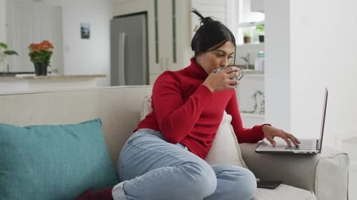 Video of happy biracial woman in hijab sitting on sofa with coffee and using laptop
