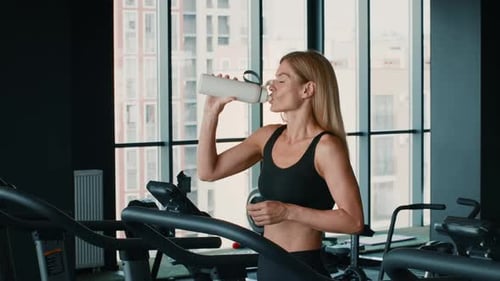 Woman Drinking Water While Exercising in a Gym With Large Windows Overlooking the City