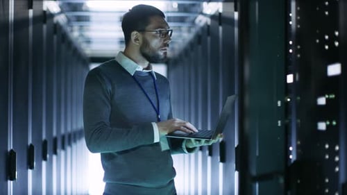 Male IT Technician Working on a Laptop Standing Before Open Server Rack Cabinet in Big Data Center.