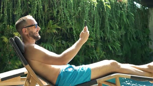 Happy young man chats on smartphone and drinks whiskey by resort pool
