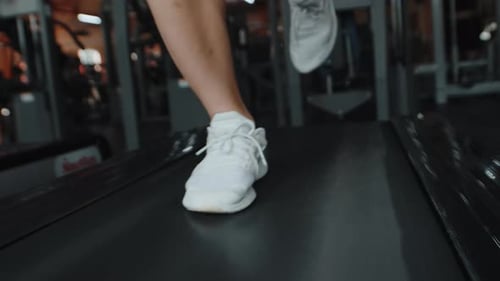 Jovem atleta em tênis brancos correndo vigorosamente em uma esteira em uma academia coberta de perto