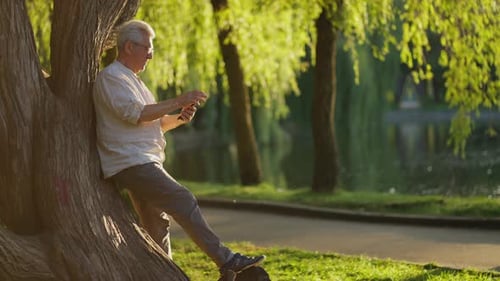 Senior Man Relaxing with Smartphone By the Lake Mature Man Enjoys Nature with Tech Concept
