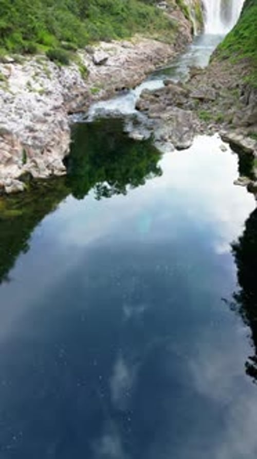 Aerial drone view a waterfall plunging into a crystal clear pool, surrounded by rocky cliffs and lus