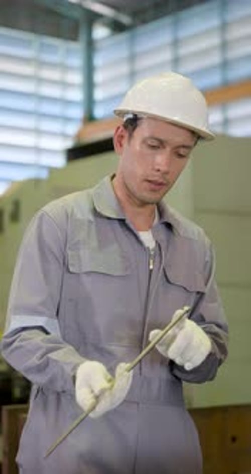 Asian adult male technician in hard hat examines long metal rod during shop floor inspection,