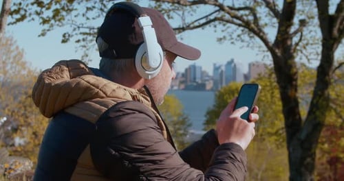 Man Listens to Music on Headphones and Looks Into a Smartphone Outdoors in Park