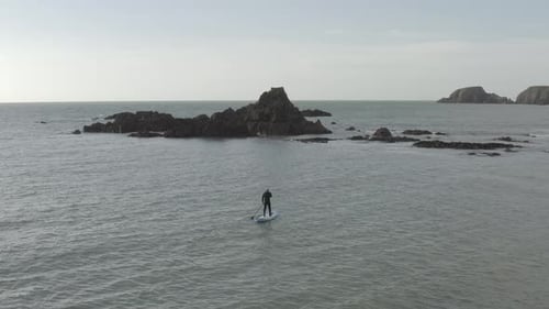 Stand up paddleboarder paddles toward rocky ocean islets on grey day