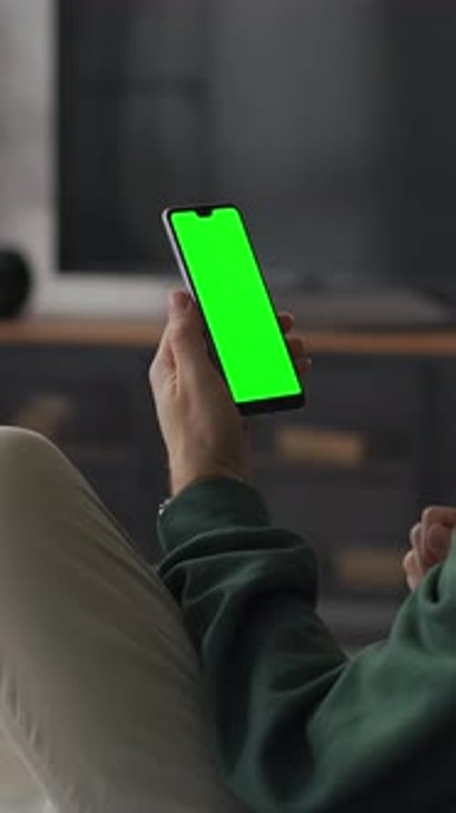 Relaxed Young Man Lounging on Sofa at Home While Holding Smartphone with Bright Green Chroma Key
