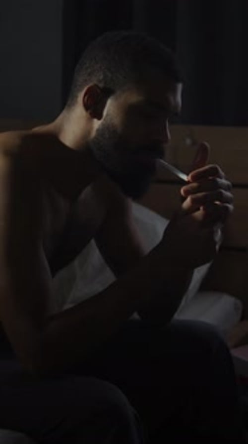 Man Smoking Cigarette on Bed in Dim Room