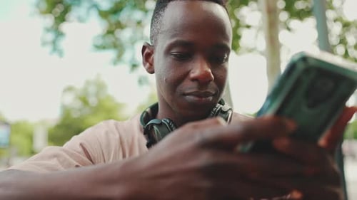 Clouse up, young student sits on bench outside of university in headphones, uses phone