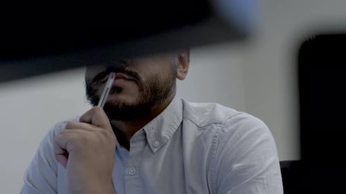 Abstract View Of Bored Employee Tapping Pen On Lips At Desk, Viewed behind two computer monitors. Lo