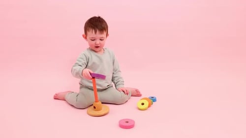 Fröhliches Kleinkind spielt mit einer Pyramide auf einem studio-rosa Hintergrund. Junge legt Holzringe an