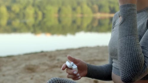 Athletic Blonde Young Woman Sits on Bank of River or Lake and Puts on Headphones to Go in for Sports