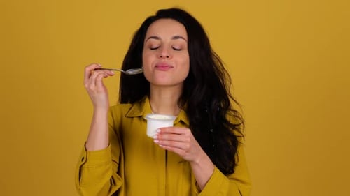 Woman Enjoying Yogurt Snack Against Yellow Backdrop