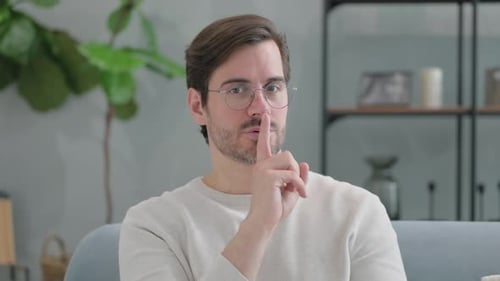Silence Please Gesture by Young Man Sitting on Sofa at Home