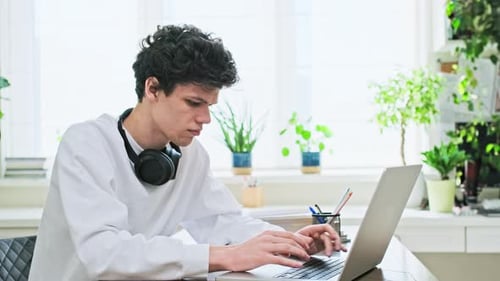 Young Male College Student Sitting at Desk at Home Typing on Laptop
