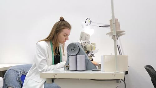 Woman Sews Grey Fabric on a Professional Sewing Machine While Sitting at Her Working Place in