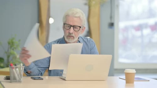 Senior Man Reviews Documents at Desk with Laptop