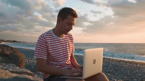 Digital Nomad Man Working with Laptop Sits on the Beach By the Sea During Sunset Male Freelancer
