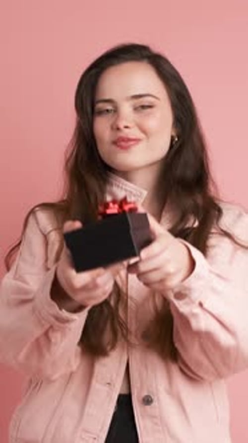 Happy Woman Giving Gift Box and Looking at Camera in Pink Studio