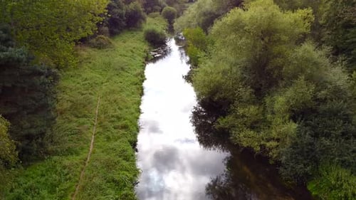 Aerial flight over the small river with the pathway on the left bank and cloud reflections on the wa