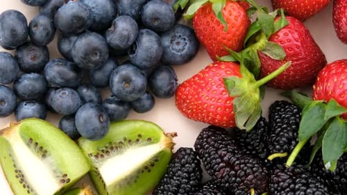Close Up Top View Of A Rotating Bowl Of Black Mulberries, Strawberries, Blueberries And Sliced Kiwi