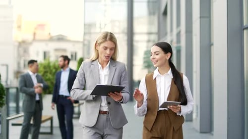 Two coworkers businesswomen walking near office building, talking about work, discussing project.