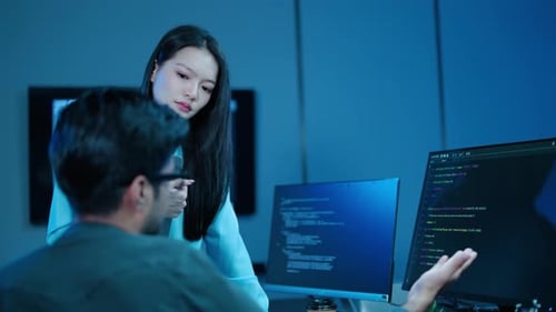 Two Asian software developers collaborating on code late at night. Male programmer points computer