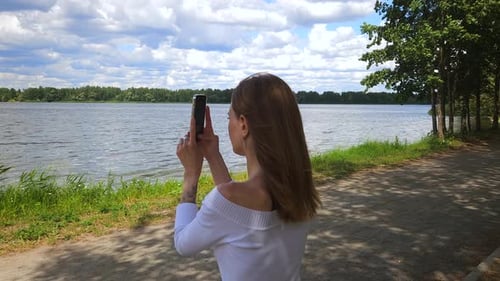 Woman Makes Video On Smartphone While Sitting On Bench On River Bank