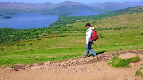 Side view of Hiker walking down a hill on the edge of a mountain ridge with lush grassy fields, hill