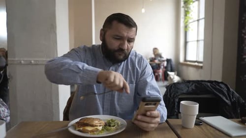Young Man Browsing Internet, Texting on Smartphone During Meal in Cafe Adult