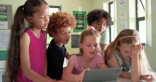 Schoolchildren leaning around tablet at classroom table, central girl touching screen to start game