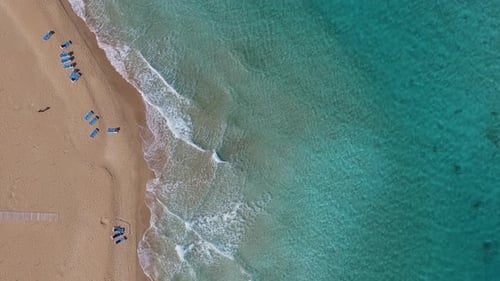 Aerial drone view of gentle waves reaching a sandy beach with turquoise water and sunbeds arranged o