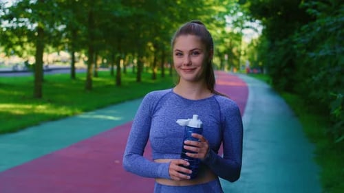 Portrait of young caucasian woman holding a bottle full of water before jogging in the park. Shot wi
