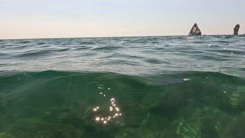 Sea Water Surface Camera Flies Over the Calm Azure Sea with Volcanic Rocky Shores on Background