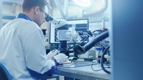 Ein Arbeiter in einer Elektronikfabrik im weißen Arbeitskittel lötet eine Leiterplatte durch eine Digitalkamera