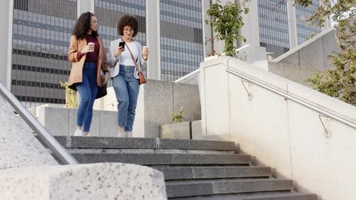 Holding coffee cups, Diverse female friends descending stairs in plaza while swiping smartphone