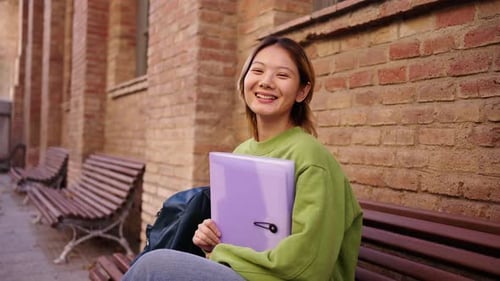 Portrait of Young Smiling Chinese Student Sitting on a Bench on Campus Looking at the Camera