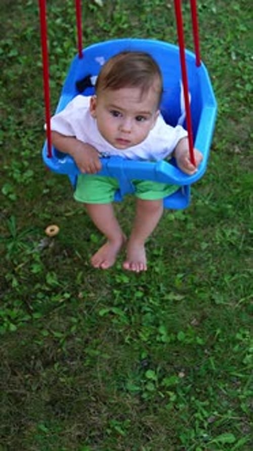 Thoughtful little baby sitting in a blue swing and swaying. Summer in the village concept.