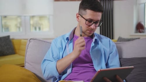 Handsome Man Sitting on Sofa Use Tablet Computer Talk and Write in a Notebook Speaking on Video Call