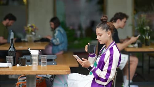 Young Woman Sitting Outdoors Cafe and Drinking Coffee Holding Smartphone