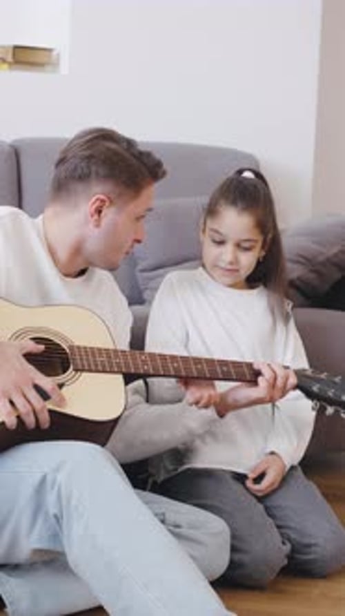 El padre enseña a su hija a tocar la guitarra durante una acogedora tarde interior en casa, creando una experiencia duradera