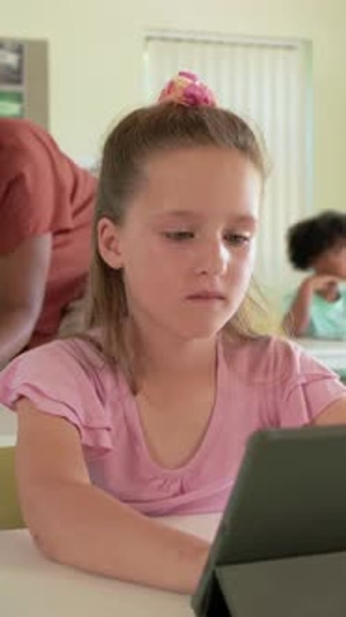 Vertical video: Tapping girl in pink top completing tablet task at school desk, teacher leaning