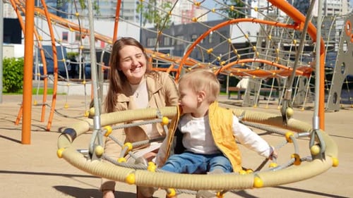 Slow motion of happy mother pushing swing with her baby son on playground