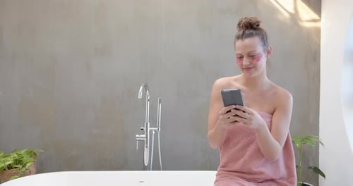 Using smartphone, woman in towel smiling and relaxing in bathroom, copy space