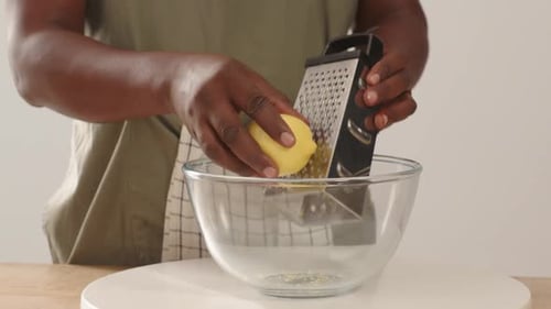 Afroamerikaner in Schürze, legt eine abgewinkelte Reibe in eine Glasschale und reibt die Zitronenschale