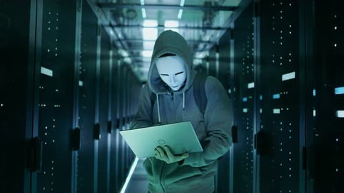 Mittelaufnahme eines maskierten Hackers in einem Hoodie, der mitten in einem Rechenzentrum voller Rack-Server steht