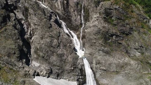 Luftaufnahme eines kaskadierenden Wasserfalls, der raue Felsklippen mit Schneeflocken hinunterfließt.