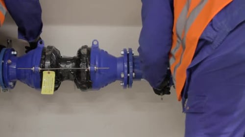 Two workers connect a hydraulic pipe connector between two pipes. Tracking shot.