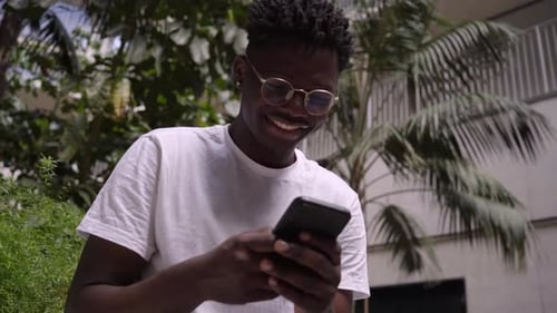 Happy Young African Using Cell Phone Sitting with Palm Trees in Background Smiling Black Man